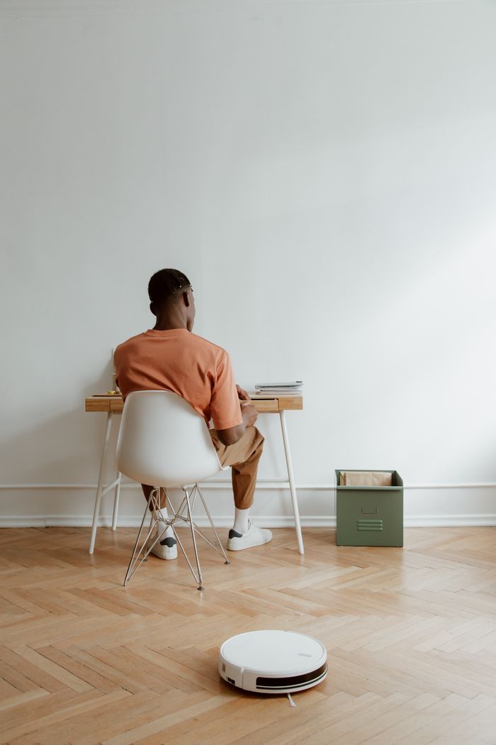 A man working at a desk in a minimalist home office with a robot vacuum cleaner on a wooden floor.